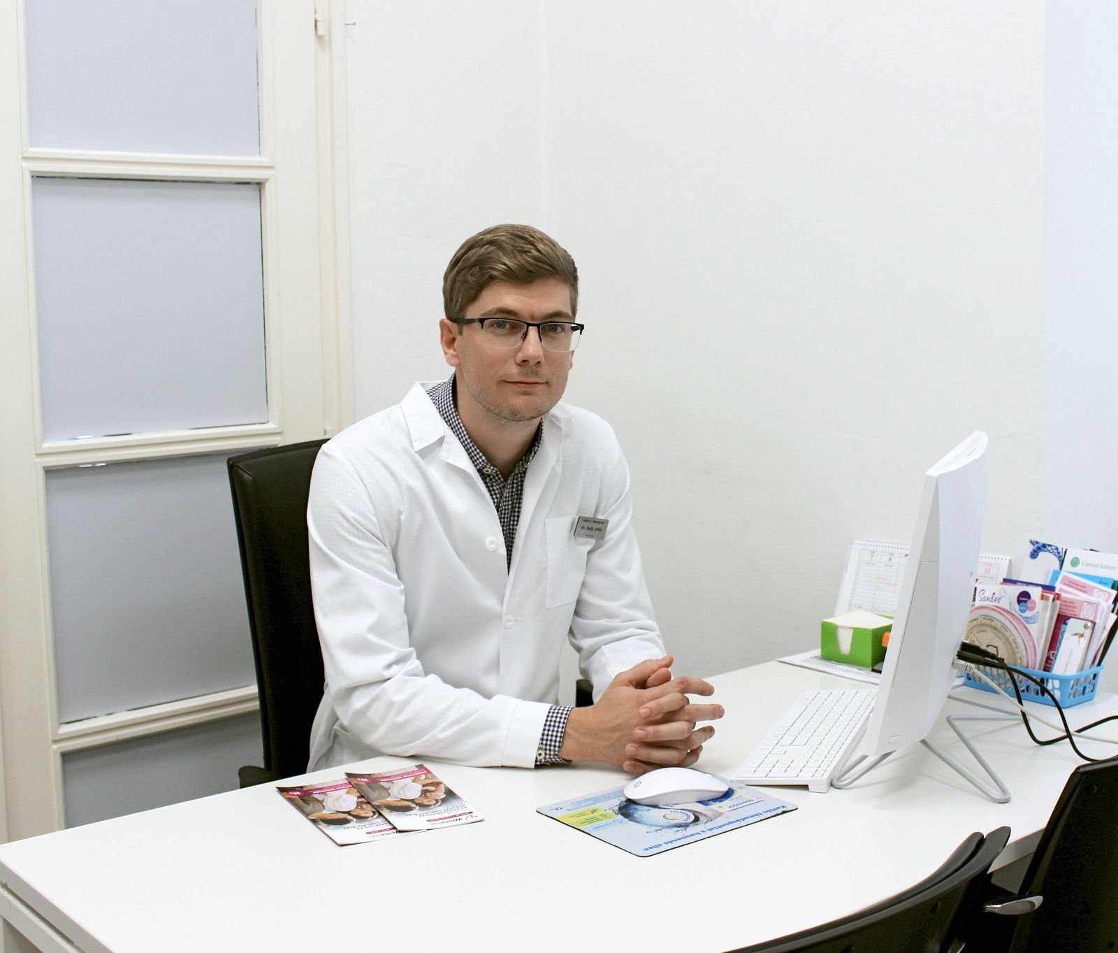 Dr. Badó Attila fehér köpenyben ül a rendelő íróasztalánál